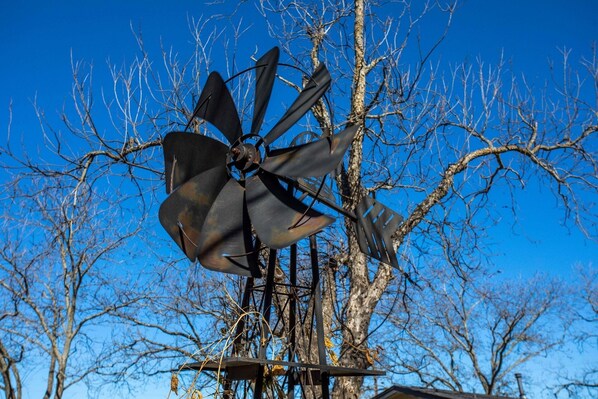 A windmill with a metal structure and blades, set against a backdrop of leafless trees and a clear blue sky.