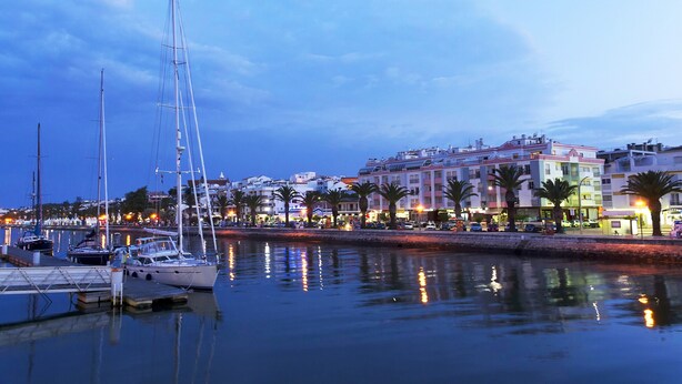 A marina with boats docked, a waterfront promenade, and buildings with balconies.