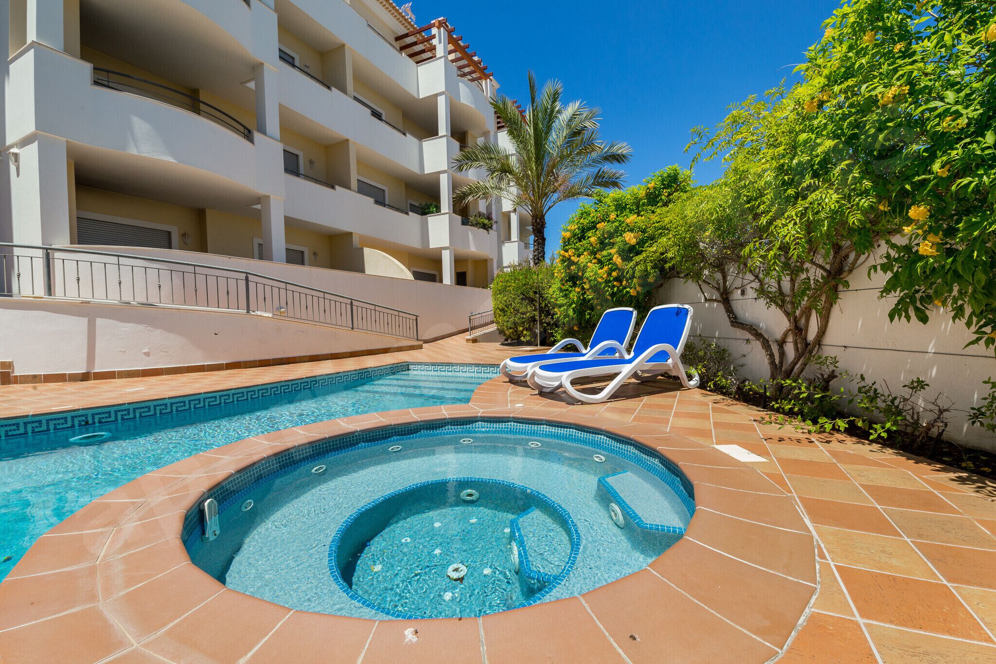 A swimming pool with a hot tub, lounge chairs, and a building with balconies.