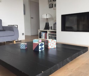 A living room with a sofa, a coffee table with geometric-patterned boxes, a bookshelf, and a mounted TV.