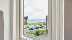 Une vue depuis une fenêtre donnant sur une rue où sont garées des voitures et sur un espace verdoyant.