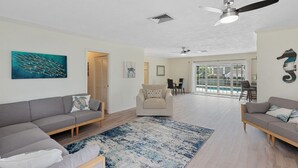 A spacious living room with a grey sofa, a beige armchair, and a blue patterned rug. There is a ceiling fan, a door leading outside, and a large window with a view of a pool. - Waterfront Piece of Paradise at Tiger Tail (Marco Island)