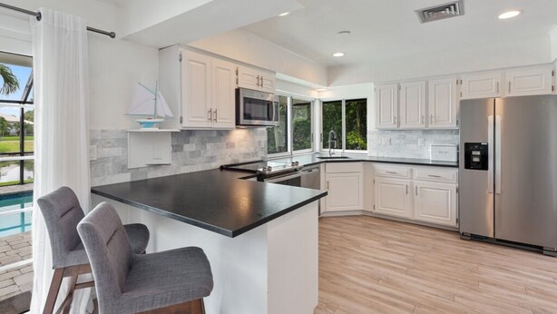 A modern kitchen with white cabinets, a large island with bar stools, a stainless steel refrigerator, and a view of a swimming pool. - Waterfront Piece of Paradise at Tiger Tail (Marco Island)