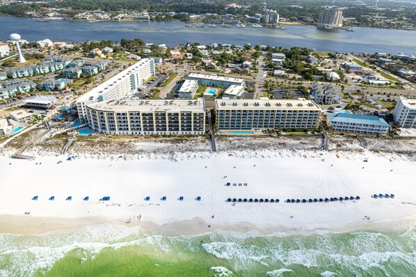 An aerial view of a coastal hotel with multiple buildings, a beach with umbrellas, and a view of the sea and cityscape.
