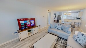 A living room with a flat-screen TV, a sofa, a coffee table, and a kitchen in the background.