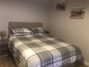 Uma cama bem arrumada com colcha xadrez, luminária de cabeceira e mesa de cabeceira pequena com luminária.