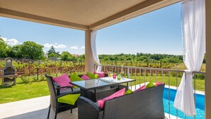 Un patio techado con mobiliario exterior, piscina y vista a un viñedo.