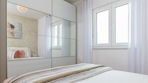 Un dormitorio con un espejo grande, una cama con ropa de cama a rayas, una ventana con cortinas blancas y un cuadro en la pared.