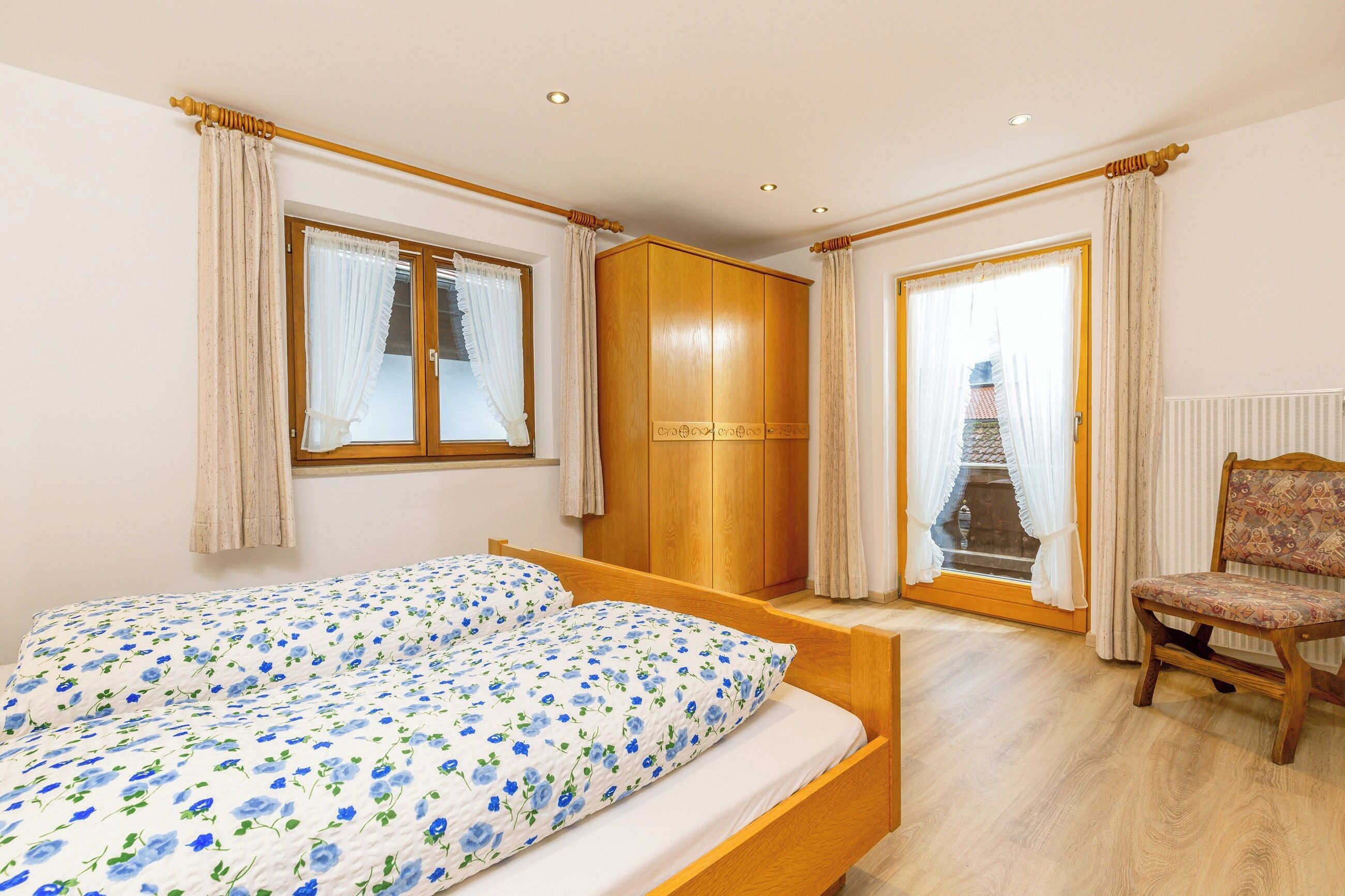 A bedroom with a bed, wooden wardrobe, chair, and a view through a window.