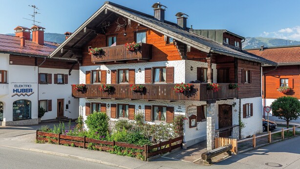 A multi-story building with wooden balconies, a sign for 'Elektra Huber', and a mountain backdrop. - Vacation apartment with balcony and mountain view, 42 sqm in a central location (Reit im Winkl)