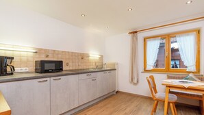 A kitchen with a microwave, sink, and wooden cabinets. There is a window with curtains and a chair at a table. - Vacation apartment with balcony and mountain view, 42 sqm in a central location (Reit im Winkl)