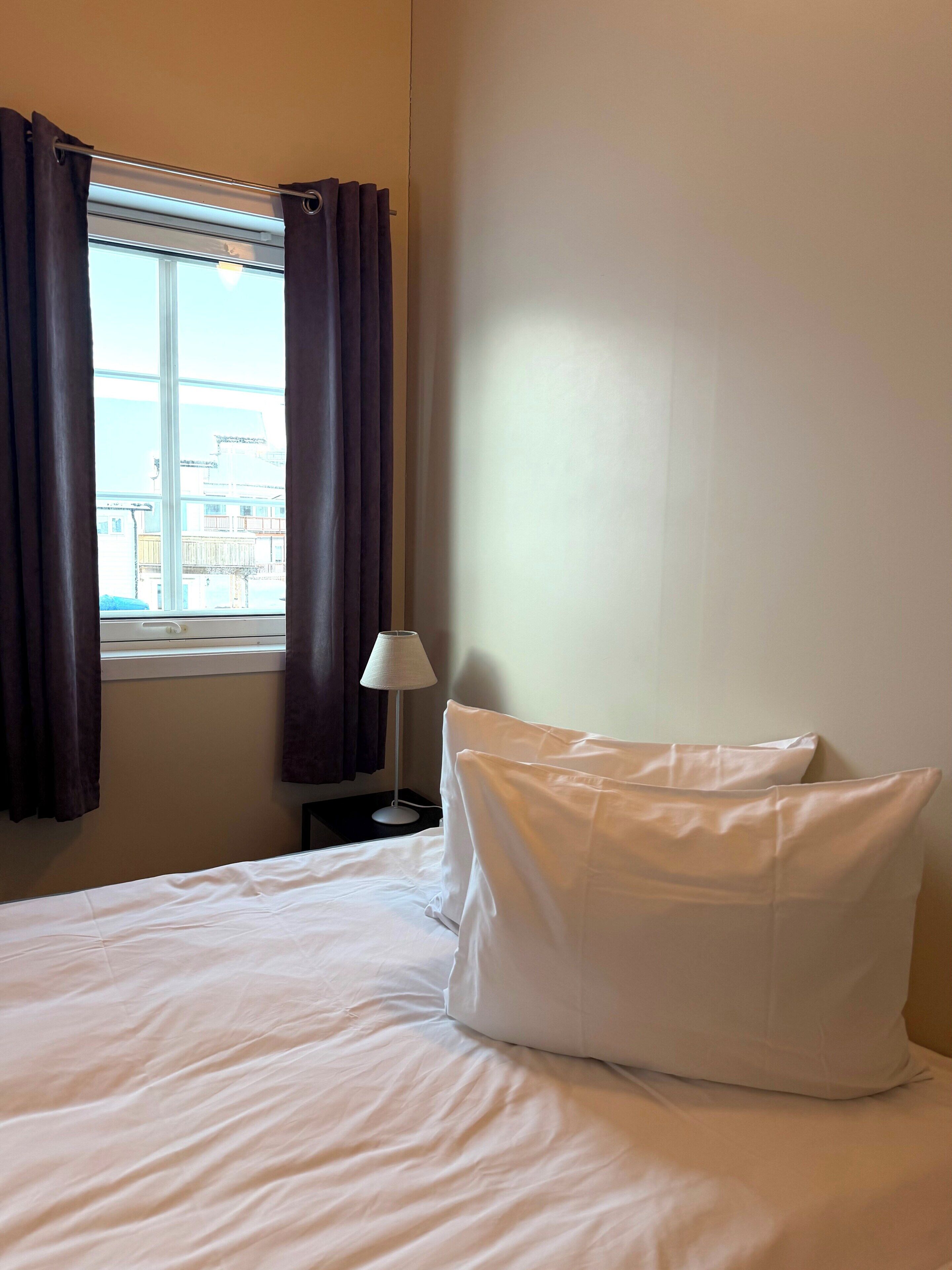 Hotelkamer met een bed, witte kussens, een nachtkastje met een lamp en een raam met gordijnen.