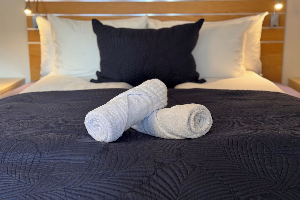 Ein Bett mit dunkler Bettdecke, zwei aufgerollte Handtücher und einem Holzkopfteil mit Leuchten.
