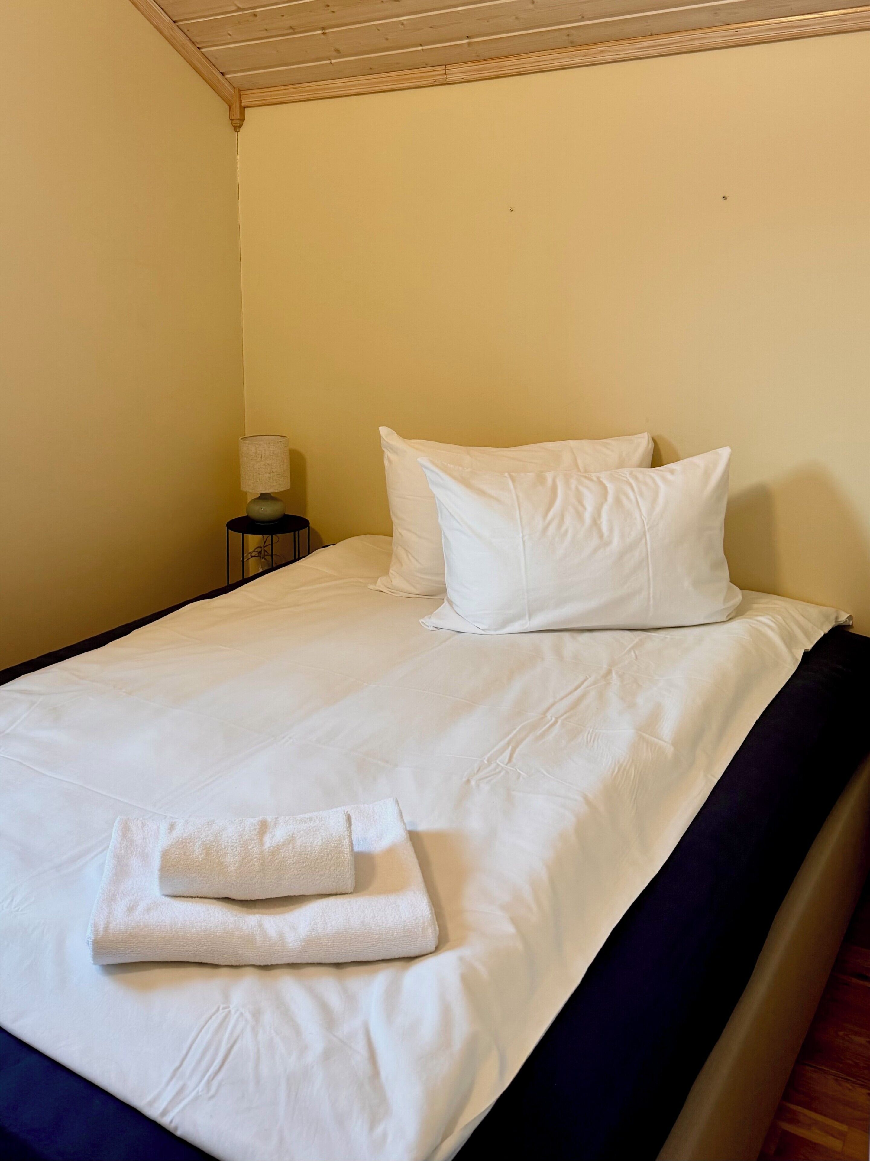 A neatly made bed with white linens, a folded towel, and a bedside lamp.
