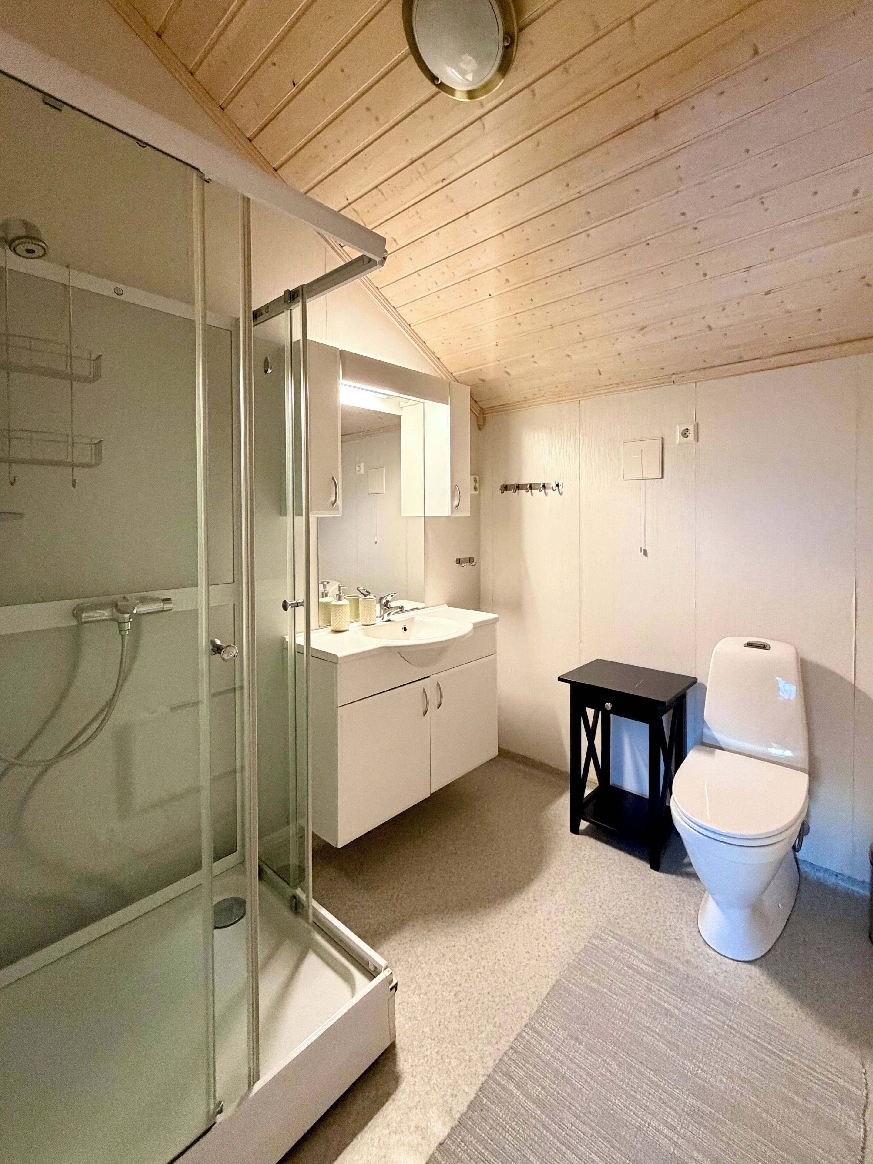 A bathroom with a glass shower enclosure, a white sink, a mirror, a toilet, and a small black table.