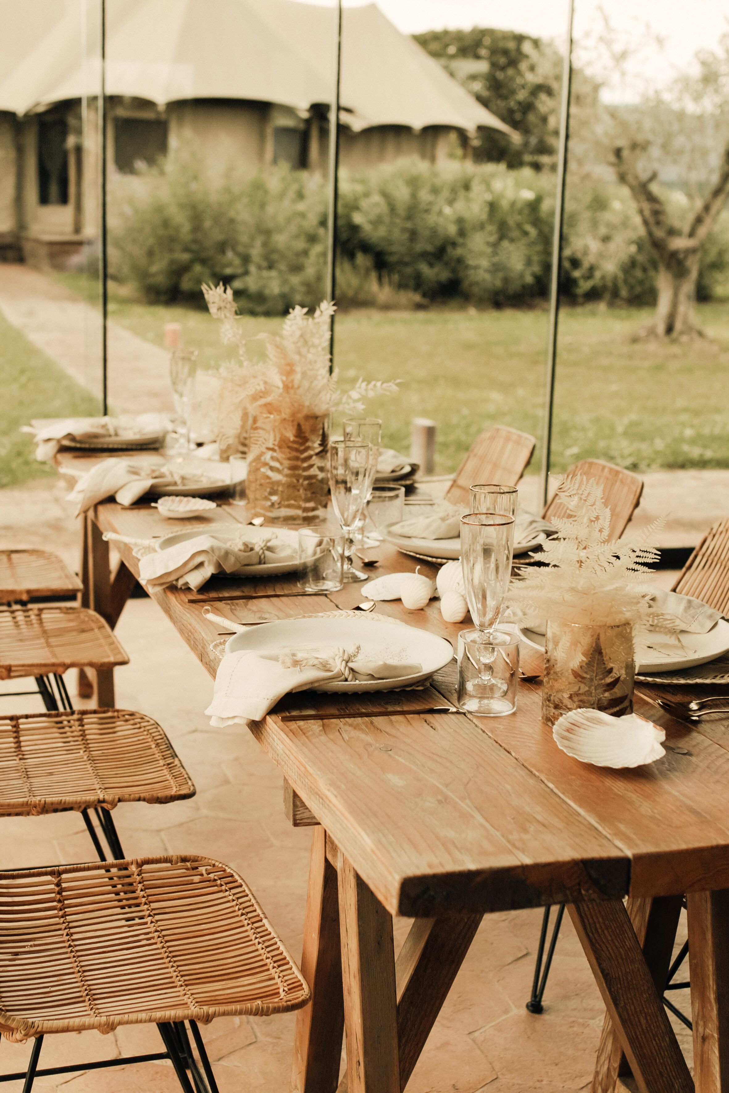 Mesa de madera lista para una comida al aire libre, con sillas de mimbre, una tienda de campaña al fondo y follaje a su alrededor.