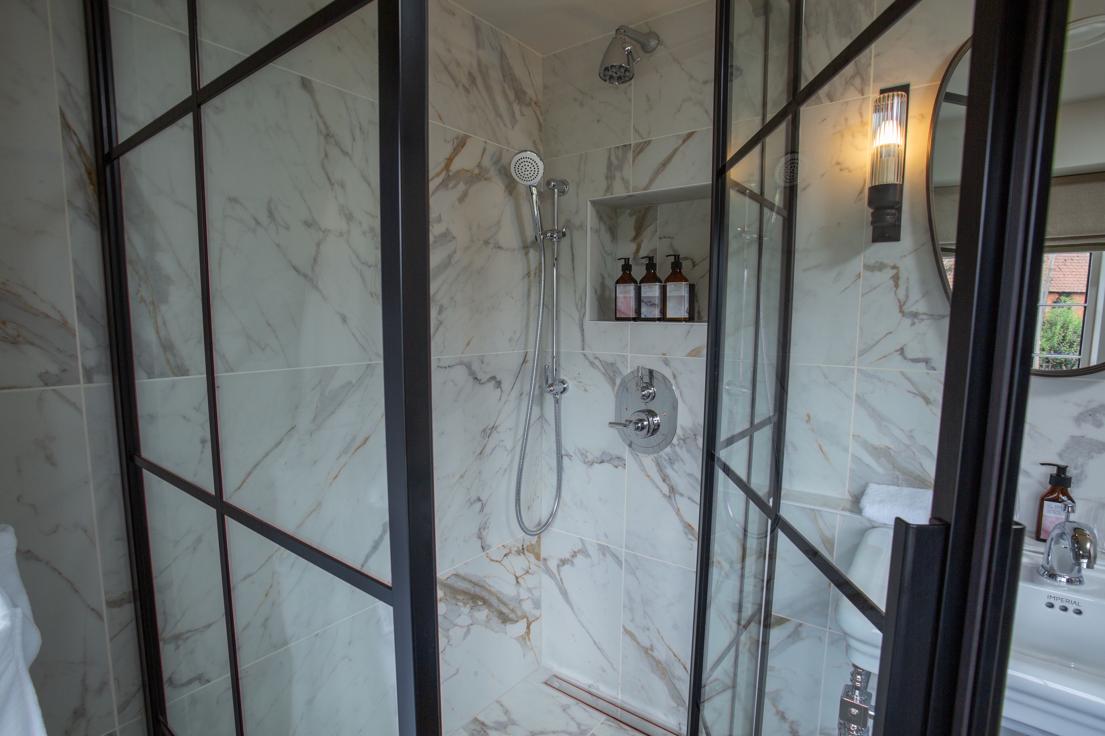 Ein modernes Badezimmer mit einer Glaskabine zur Dusche, Marmorfliesen, einem Waschtisch mit Spiegel und einem Ablagebereich für Toilettenartikel.