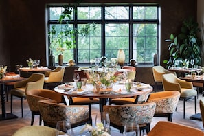 A dining area with round tables, chairs, and a large window with greenery outside. - The Falcon Hotel (Northampton)