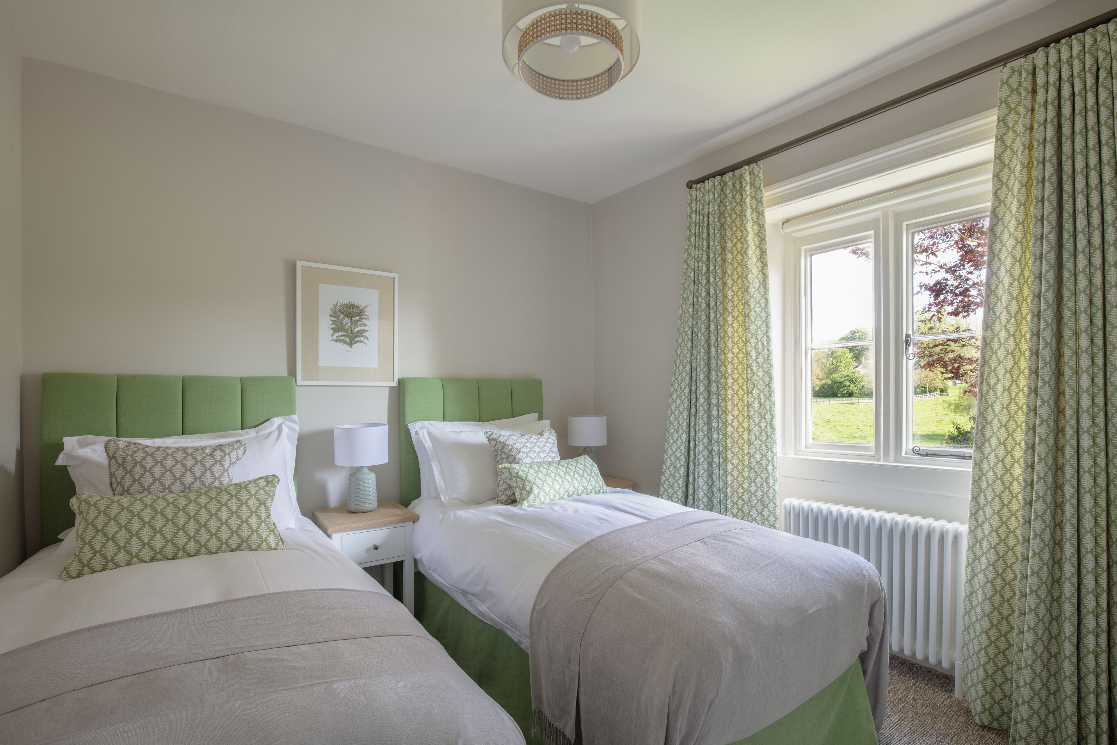 A bedroom with two beds, a green headboard, a framed botanical print, a bedside table with a lamp, and a window with curtains.