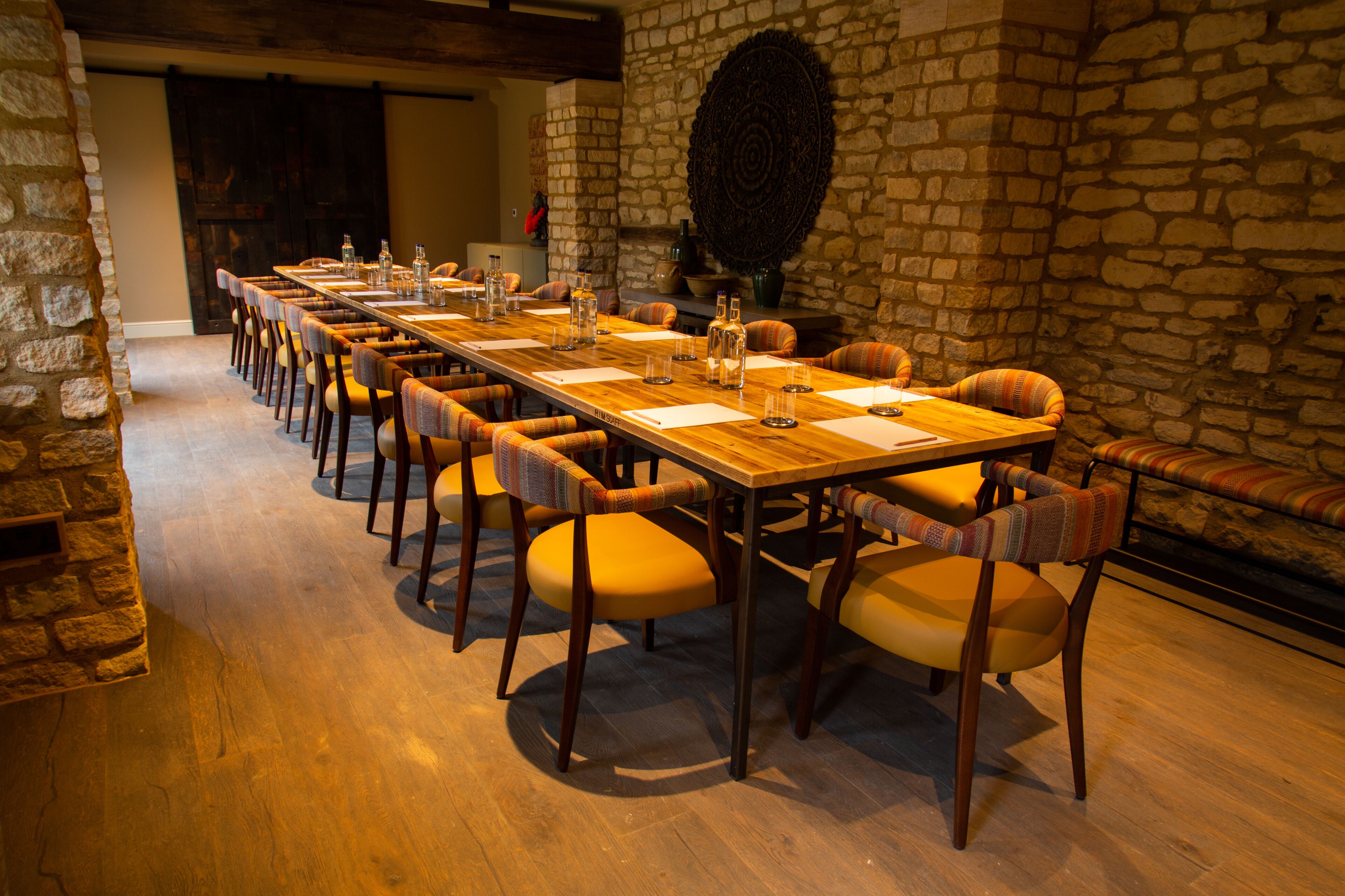 A long wooden table with chairs, set against a stone wall with a circular decorative piece.