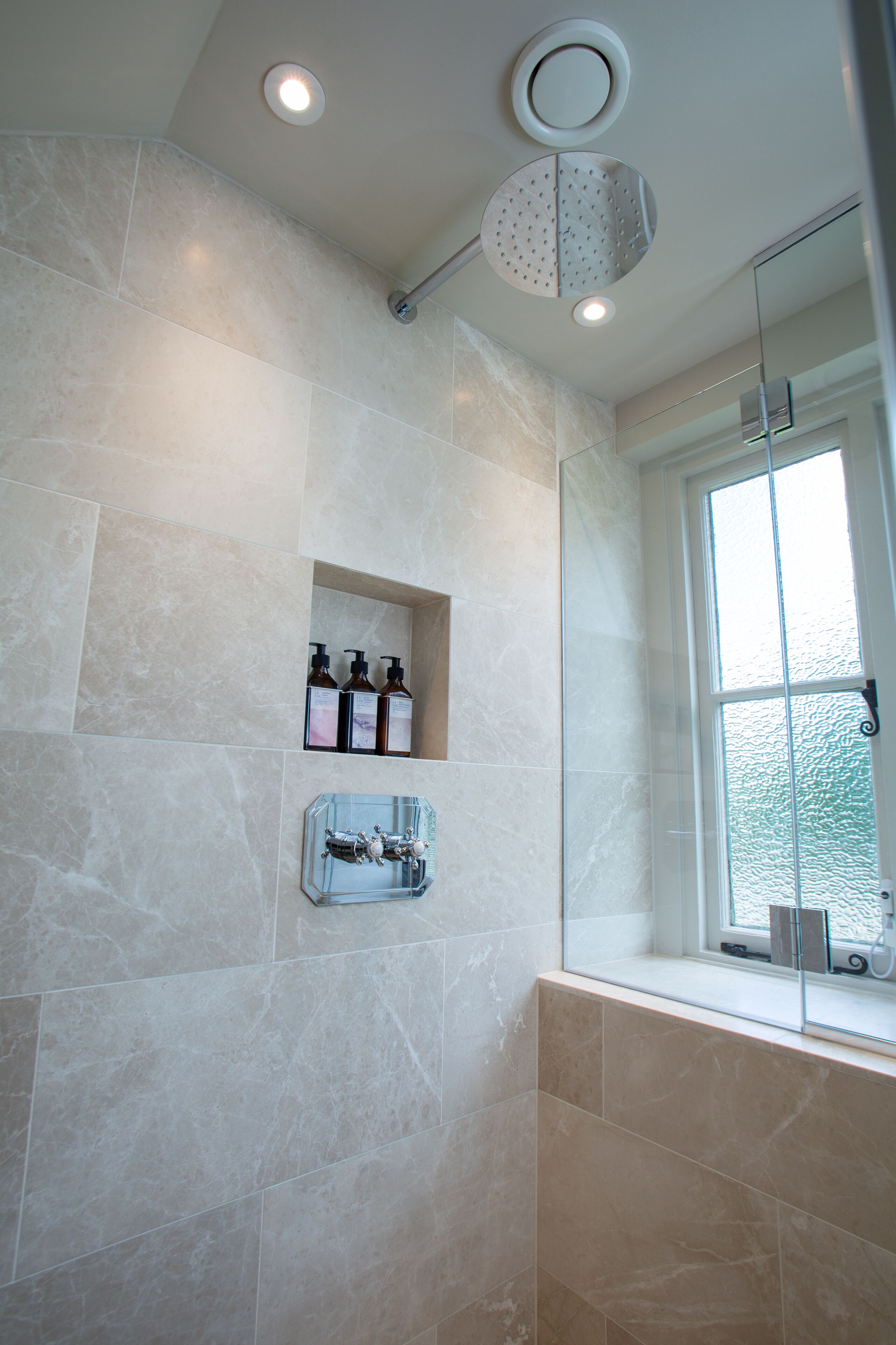 A bathroom with beige tiles, a glass shower enclosure, a window with a view, and a shelf with toiletries.