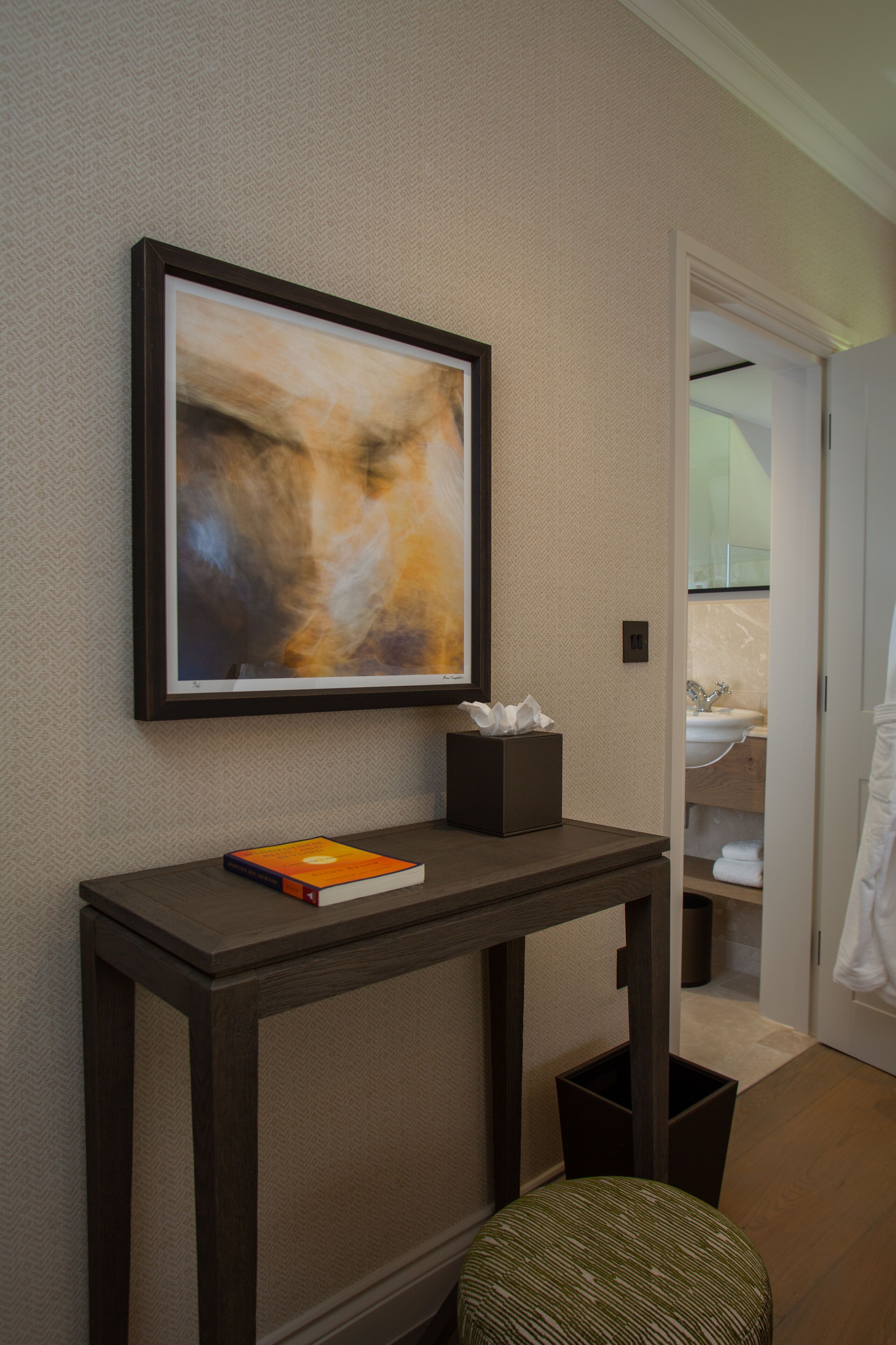 A modern bathroom with a framed abstract painting, a small table with a book and tissues, and a visible sink area.