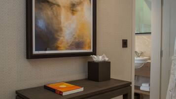 A modern bathroom with a framed abstract painting, a small table with a book and tissues, and a visible sink area.