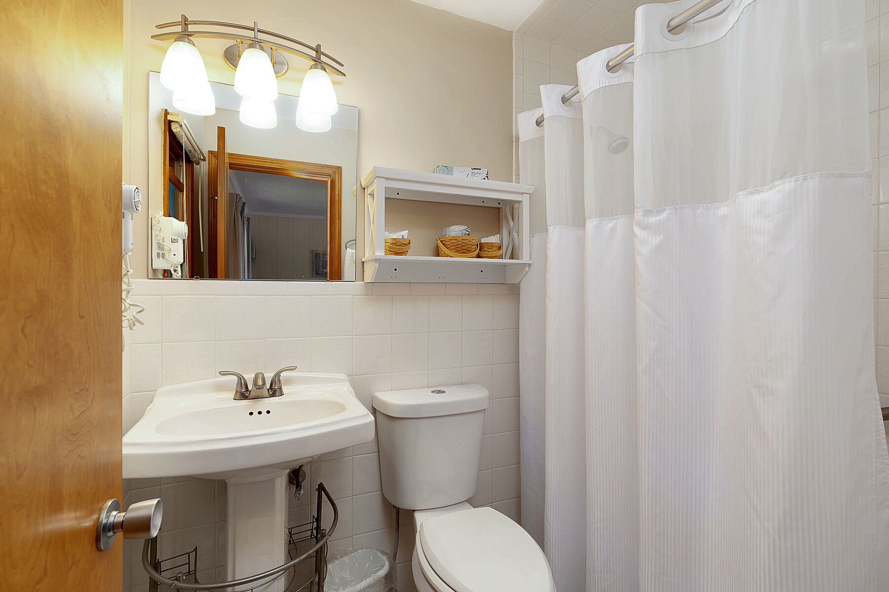 A bathroom with a sink, mirror, and toilet. There is a shelf above the toilet with baskets.
