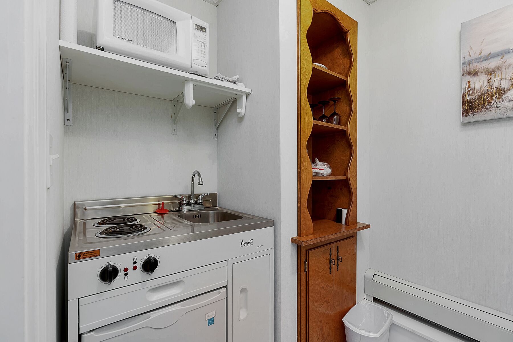 A compact kitchen with a stove, sink, and microwave. There is a wooden cabinet with shelves and a trash bin.