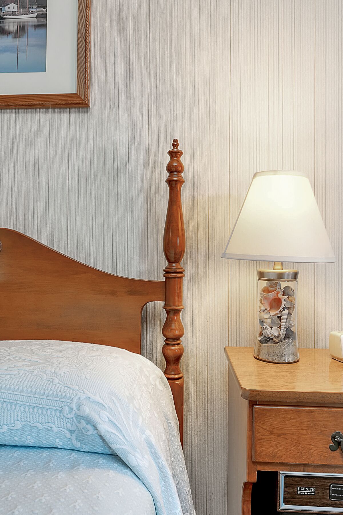 A neatly made bed with a wooden headboard, a bedside lamp with a decorative base, and a framed picture on the wall.