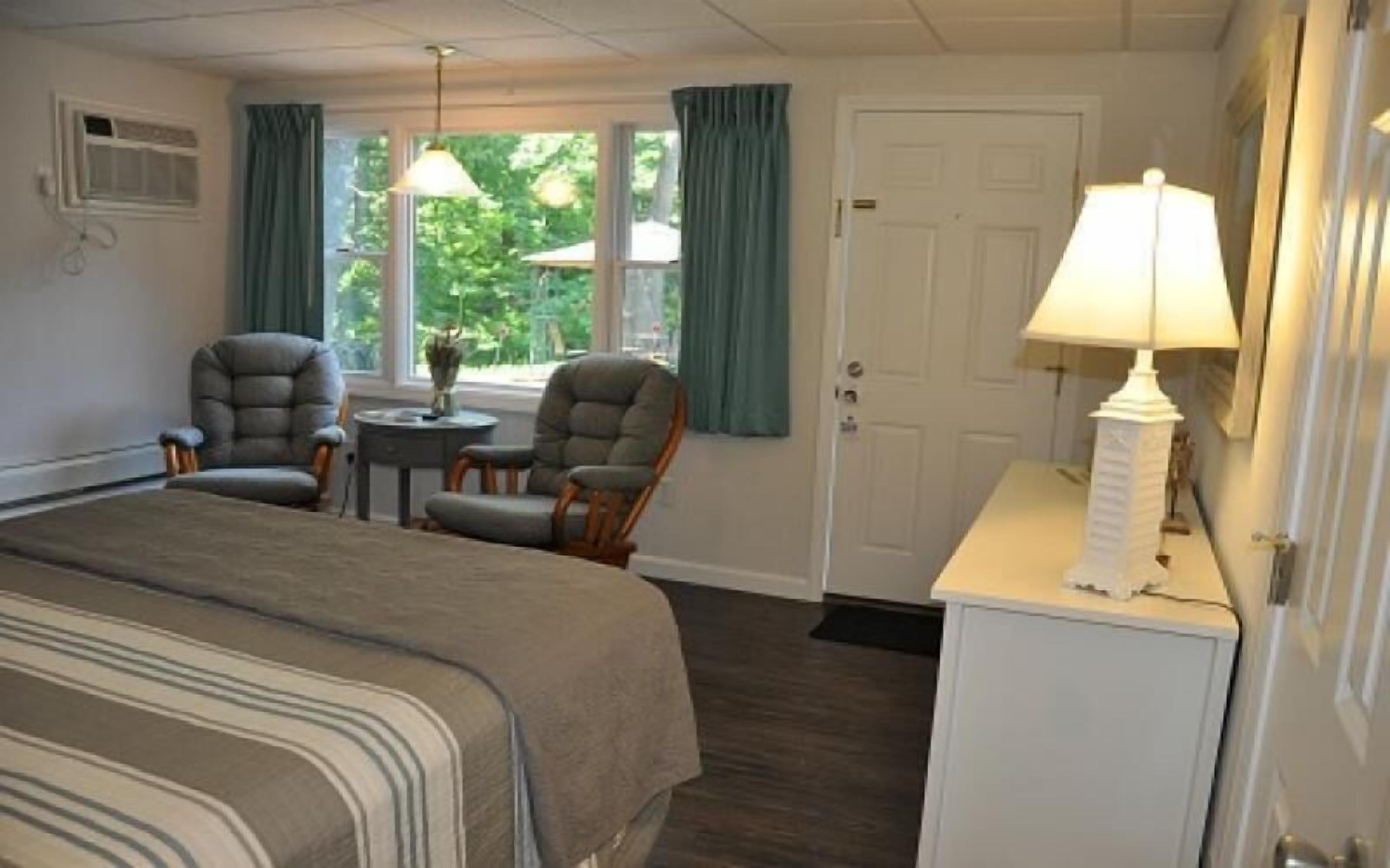A hotel room with a bed, two armchairs, a small table with a lamp, and a view of greenery outside.