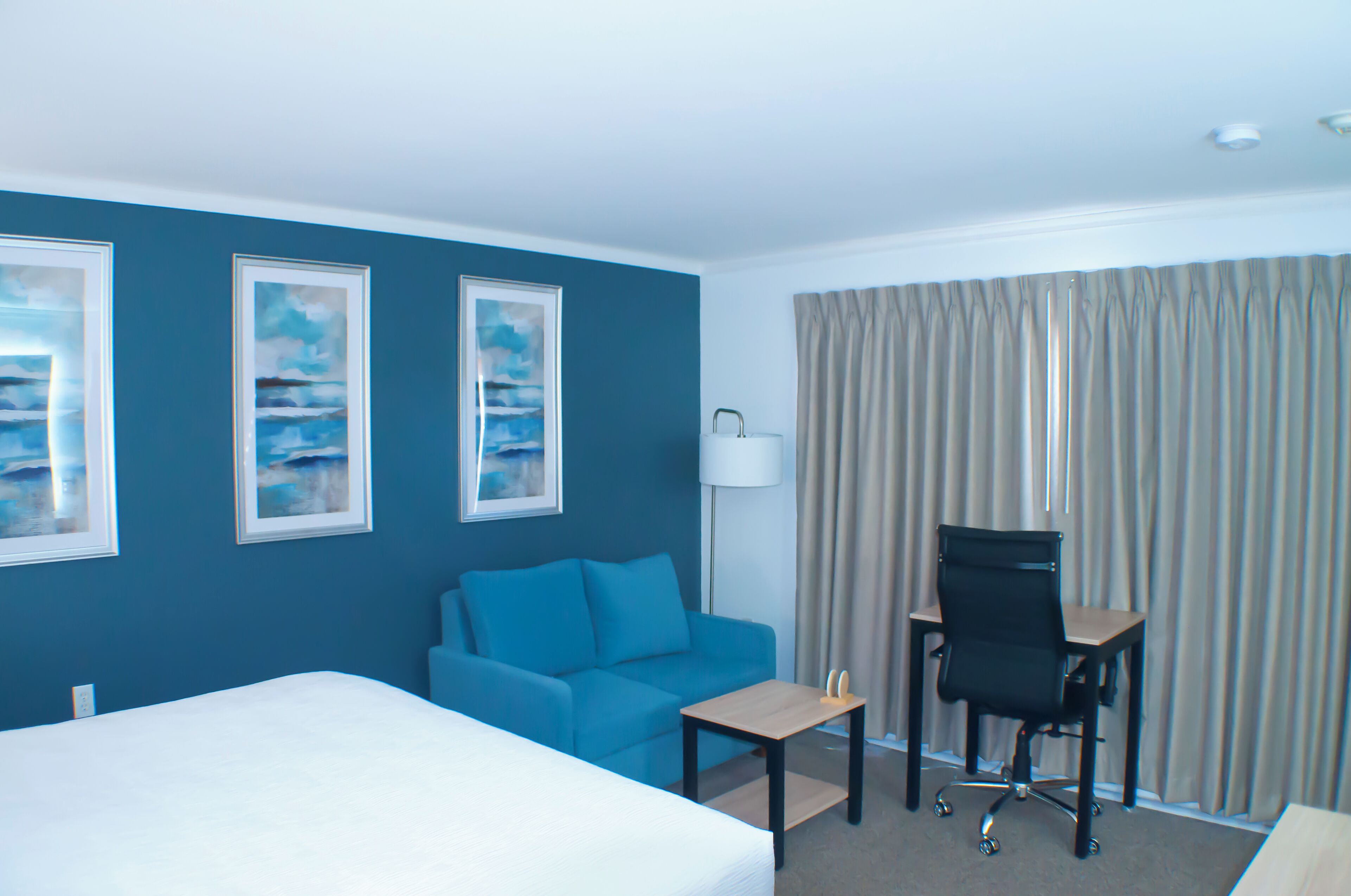 A hotel room with a bed, a blue sofa, a desk, and a chair. There are two framed pictures on the wall and a lamp on the desk.