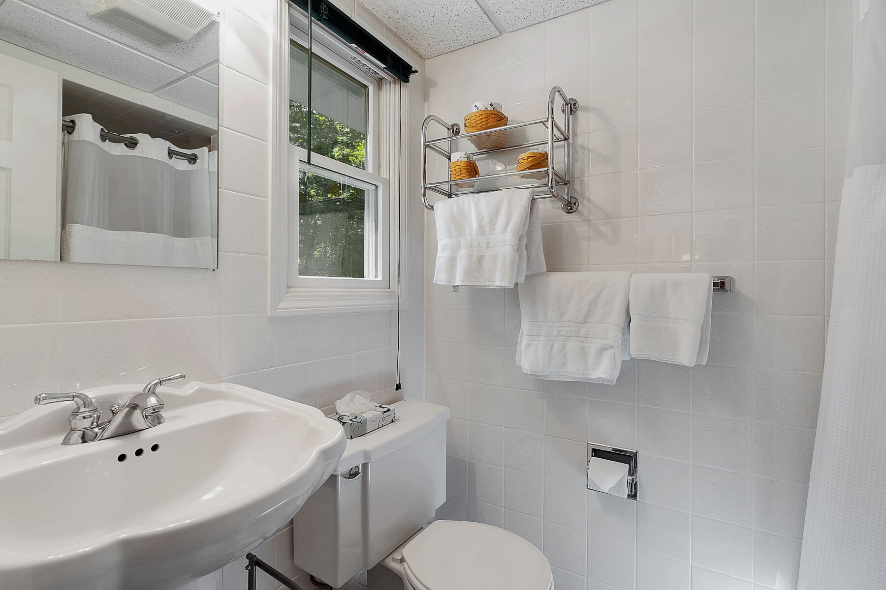 A bathroom with a sink, toilet, and a window. There are towels hanging on a rack and a basket on the wall.
