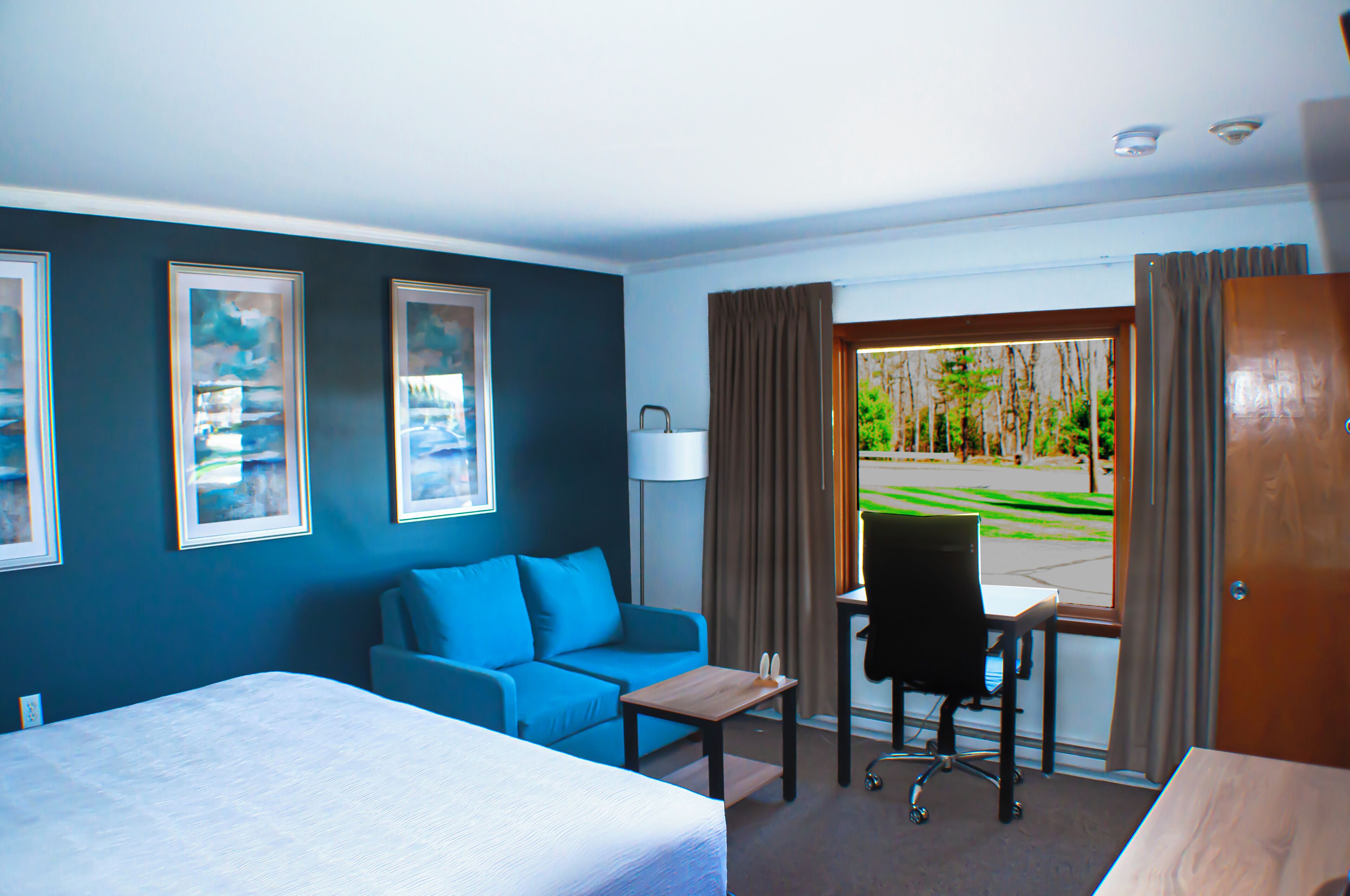 A hotel room with a bed, a blue sofa, a small table, a chair, and a window with curtains.