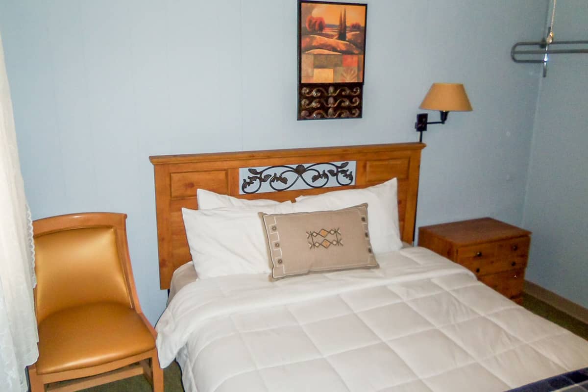Un lit bien fait, avec une couette blanche matelassée, une tête de lit en bois ornée de motifs décoratifs, un oreiller beige et une petite table de chevet en bois avec une lampe.