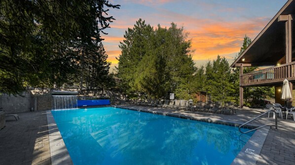 A swimming pool with a waterfall feature, surrounded by trees and a building with outdoor seating.