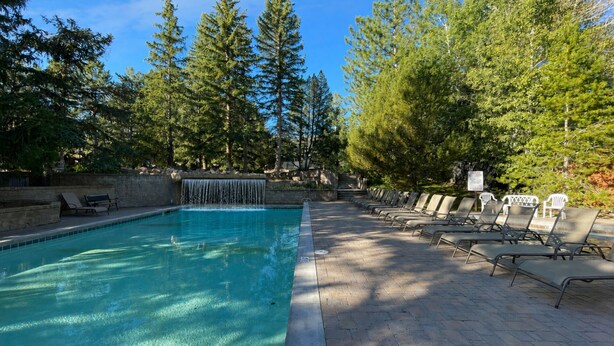 A swimming pool with a waterfall feature, surrounded by trees and lounge chairs. - Family Mountain Oasis: Pools, Hot Tubs & Summer Fun! (Park City)