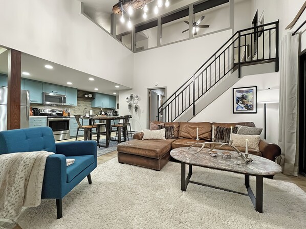 Un espacio habitable amplio con un sofá azul, un sofá de cuero marrón y una mesa de centro redonda. Una escalera con barandilla negra conduce a un nivel superior.