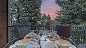 Ein Esstisch, gedeckt für sechs Personen, mit klarem Glas und Geschirr, befindet sich auf einem Balkon mit Blick auf hohe Nadelbäume und einen Sonnenuntergangshimmel.