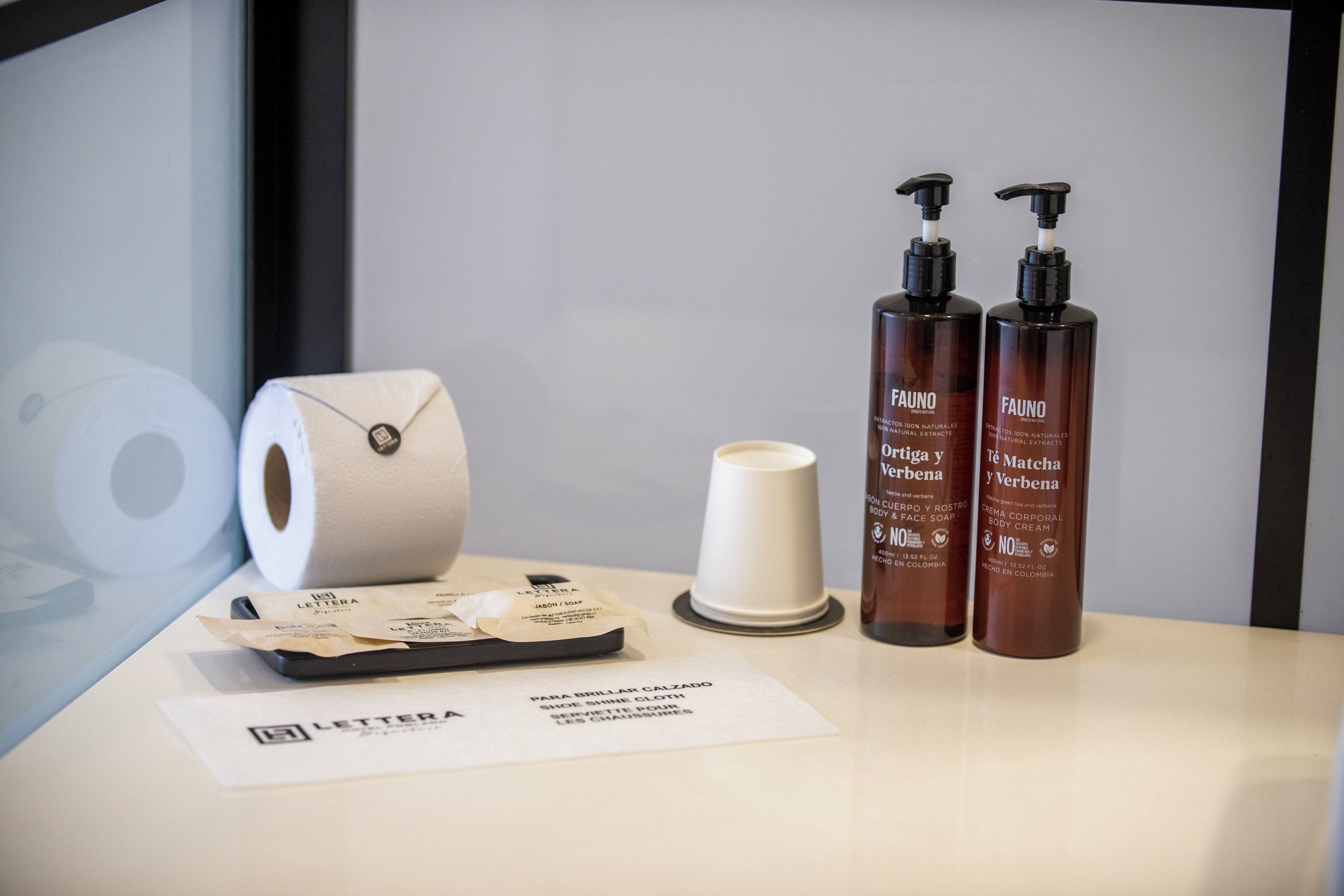 A hotel room with a roll of toilet paper, a cup, and two bottles of Fauno body cream on a table.