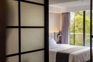 A hotel room with a bed, a nightstand, a lamp, and a view of trees through large windows.