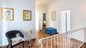 Un couloir avec un fauteuil en osier, une petite table ronde et une nappe bleue. Des œuvres d’art encadrées ornent les murs, et une rambarde blanche se trouve au premier plan.