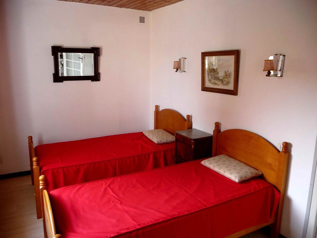 Une chambre avec deux lits en bois, chacun recouvert d’une couverture rouge et agrémenté d’un oreiller. On y trouve des lampes fixées au mur et un tableau encadré.