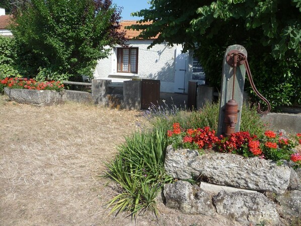 A rustic stone well with a metal pump, surrounded by red flowers and green plants, with a white building in the background. - House in Barbâtre on the Île de Noirmoutier. Wi-Fi La Cheminée 56 m² -------- (Barbâtre)