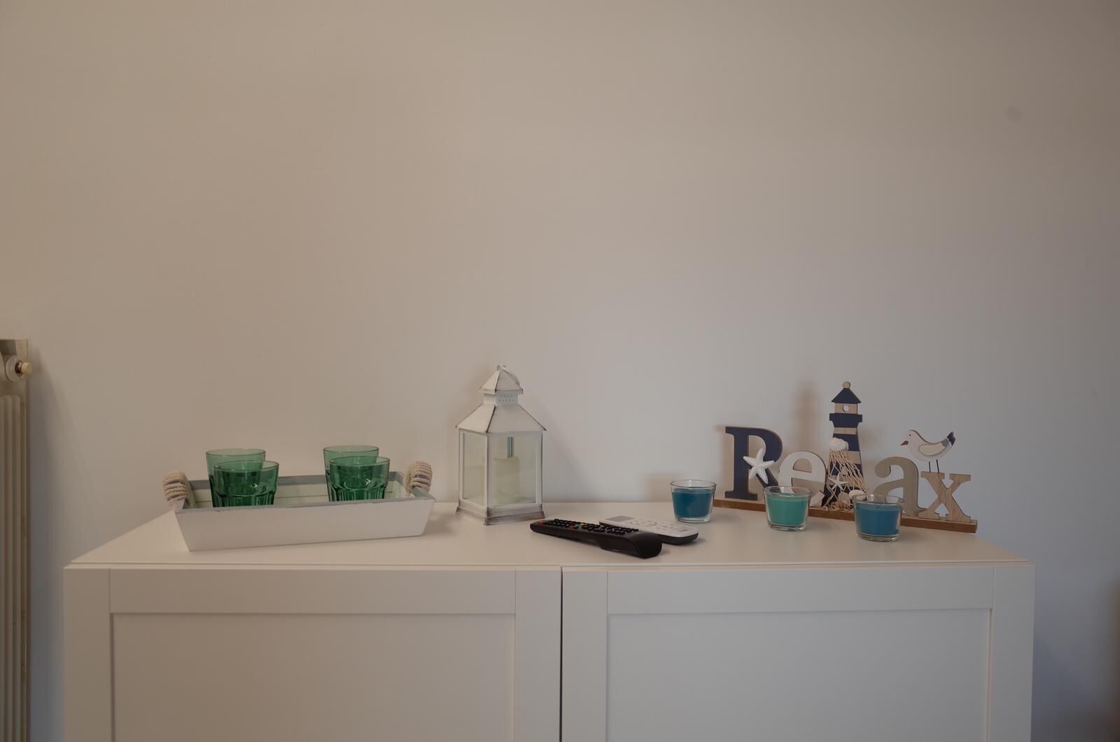 Un meuble blanc contenant un plateau sur lequel sont posés des verres verts, une lanterne blanche et une télécommande. On y trouve également des objets décoratifs, notamment un phare et une mouette.