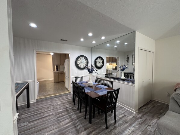 A dining area with a wooden table and chairs, a wall clock, and a kitchen in the background. - Family Ready: POOL :3BR/2BA Beach Cottage: Less than a block from Beach: WIFI (Panama City Beach)