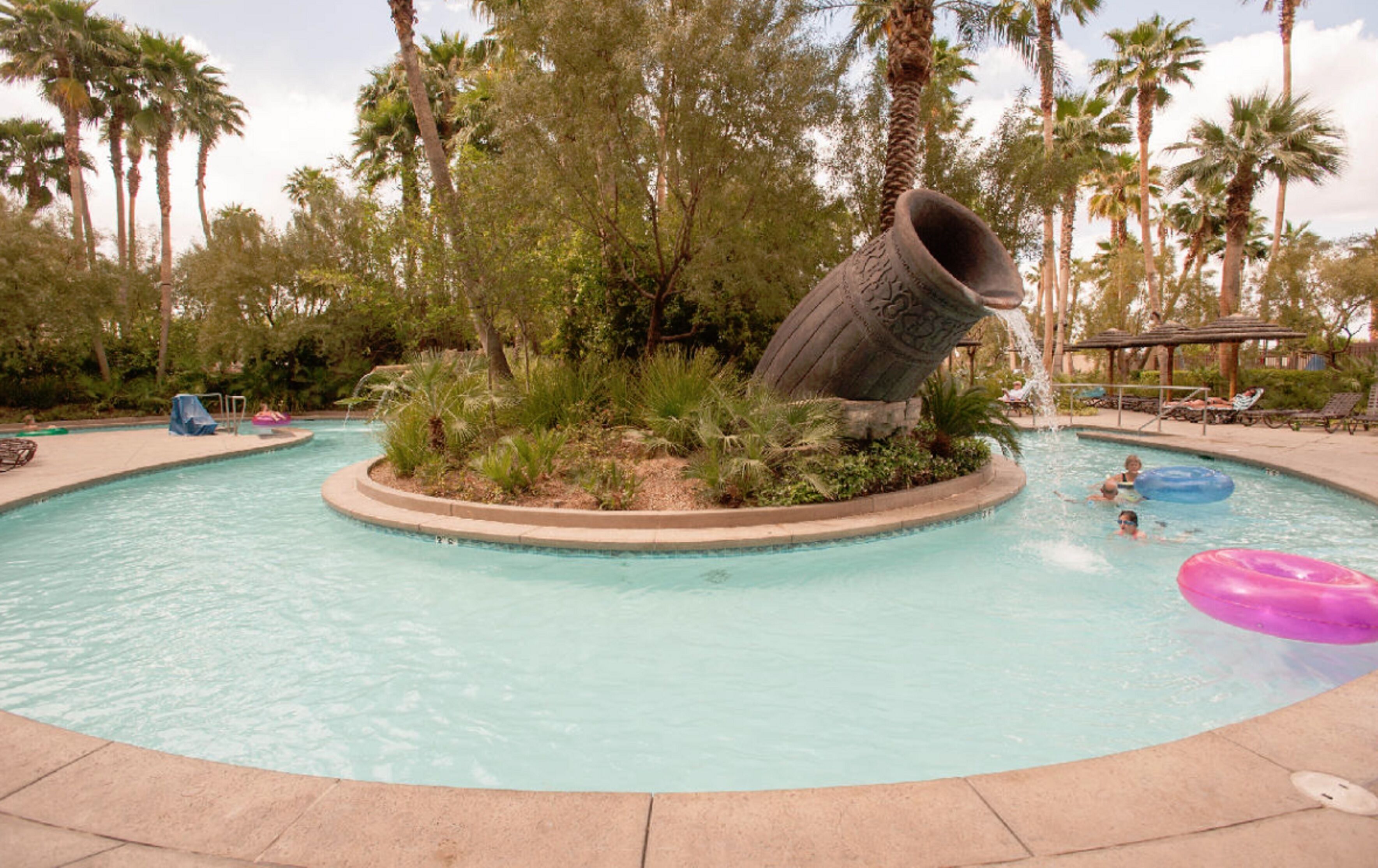 A swimming pool with a central water feature, surrounded by palm trees and lounge chairs.
