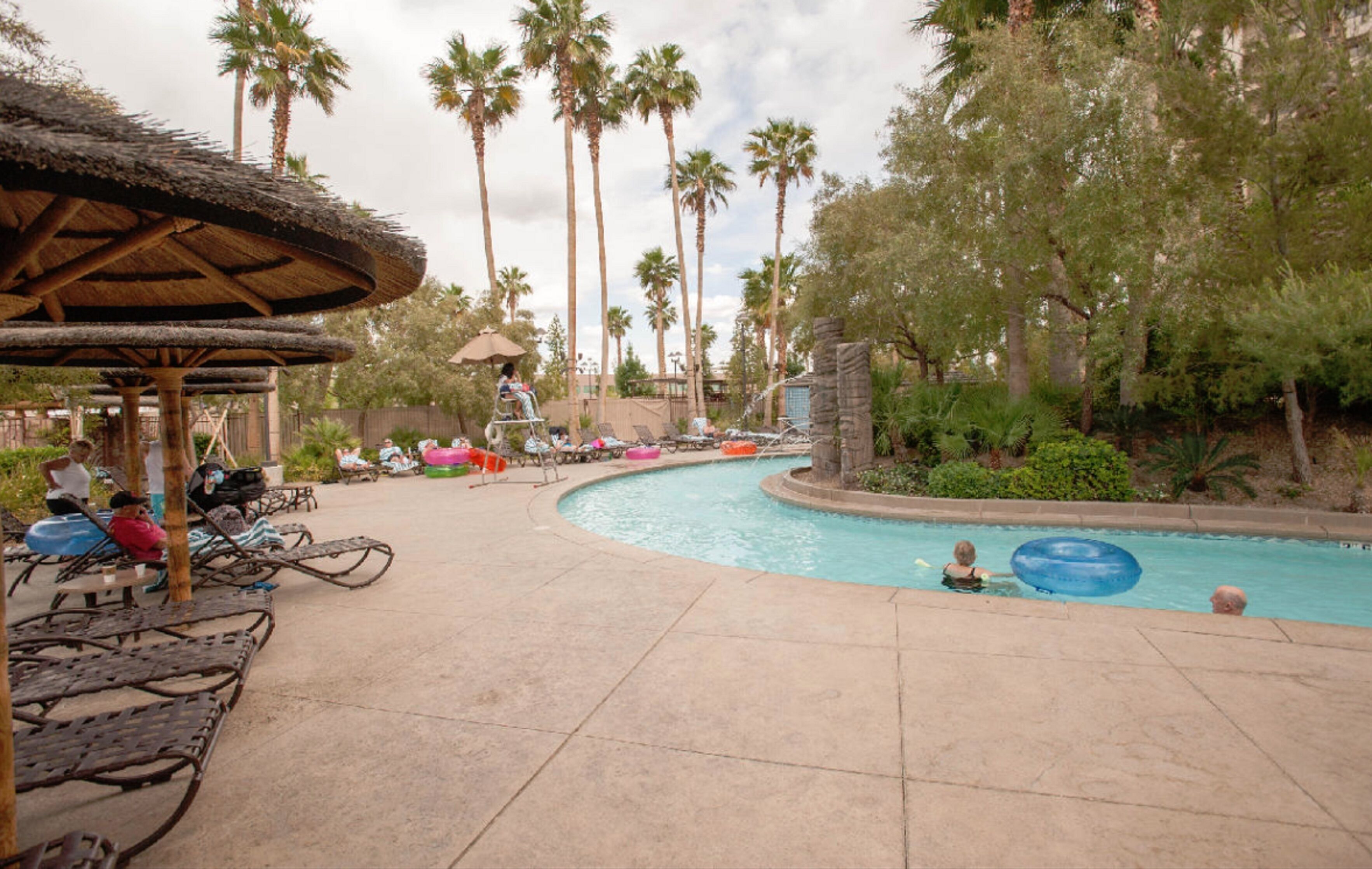 A pool area with lounge chairs, a thatched-roof structure, palm trees, and a curved pool.