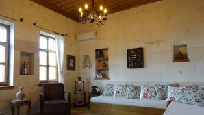 A cozy living room with a wooden ceiling, a sofa with patterned cushions, a vintage armchair, a small side table, a rug, and a wall-mounted air conditioner. - 3-Bedroom Stone House with a View (Nevşehir)