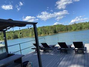 A deck with lounge chairs and a table, overlooking a lake with a forested shoreline. - Deep Water Dock -two story! Bring your boat! Cozy pet friendly cottage (Fair Play)